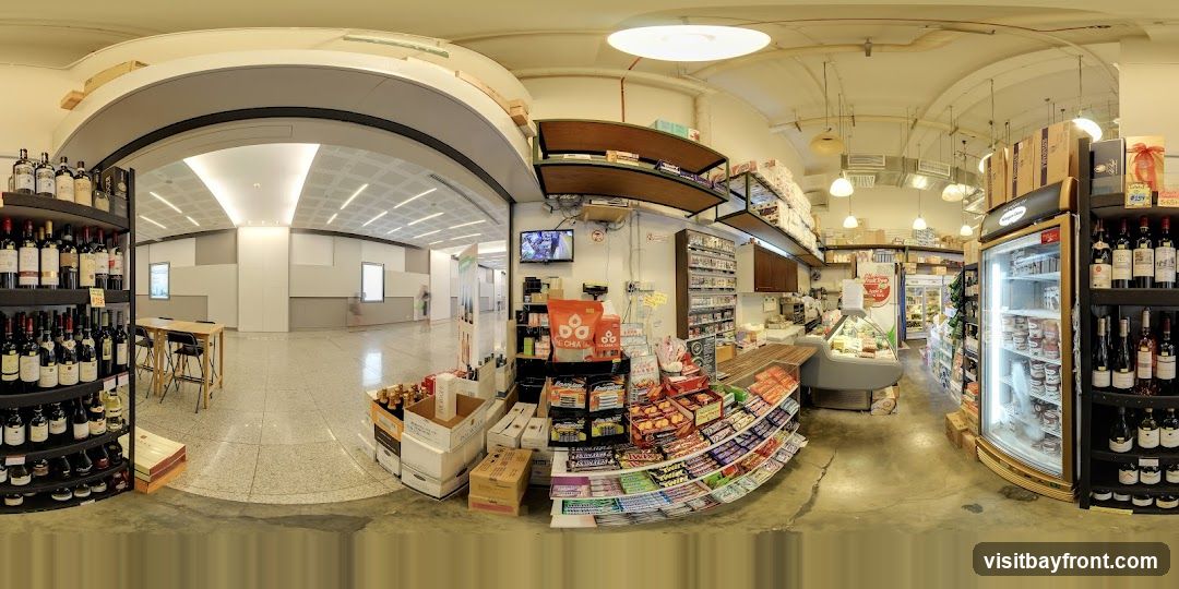 A panoramic view of the interior of a grocery store. Shelves are stocked with wine bottles, snacks, and other food items. Refrigerated display cases show various food products. A television screen is mounted on the wall. The store appears to be well-lit with overhead lighting.