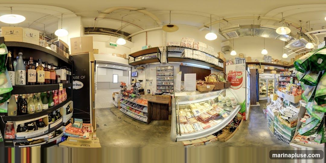 The image shows the interior of a convenience store with shelves stocked with various products. On the left, there are shelves filled with bottles of alcohol, likely sake and wine, along with some packaged goods. In the center, a counter area is visible with a cash register and more shelves displaying snacks and drinks. A refrigerated display case filled with food items is prominent on the right. The store appears to have a wide selection of imported goods and fresh produce.
