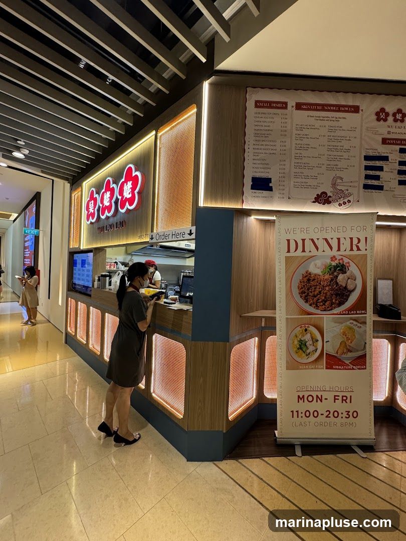 A woman stands at a counter in a restaurant, ordering food. The restaurant has a prominent sign with Chinese characters and the name "WU LAO LAO". A menu board is visible above the counter, listing small dishes and noodle bowls. A banner on the right announces "WE'RE OPENED FOR DINNER!" and displays opening hours.