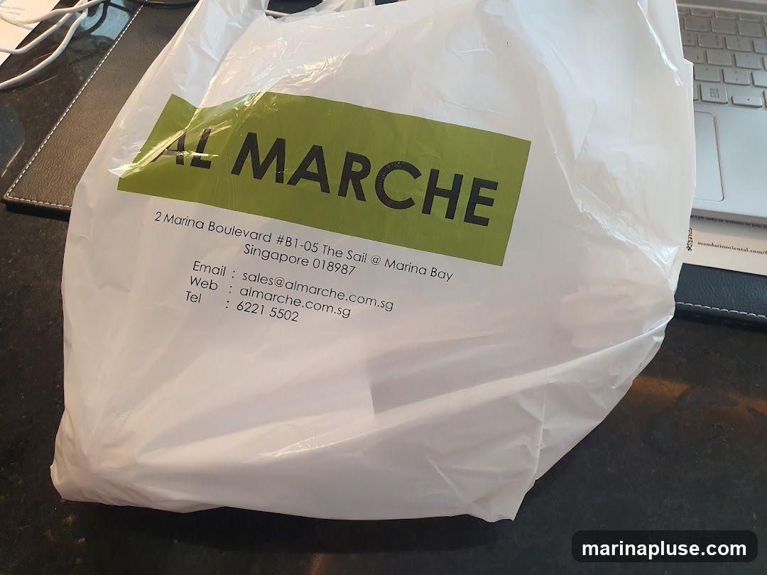 A white plastic shopping bag with the Al Marche logo and contact information printed on it. The bag is placed on a dark surface, possibly a desk, next to a laptop keyboard.