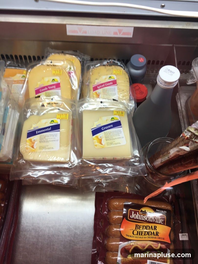 A refrigerated display case filled with various types of cheese and packaged sausages. Visible cheeses include Gouda Young, Spiced Gouda, Emmental, and Gruyere. A package of Johnsonville Beddar Cheddar Smoked Sausage & Cheddar Cheese is also present. Several bottles and containers are visible in the background.