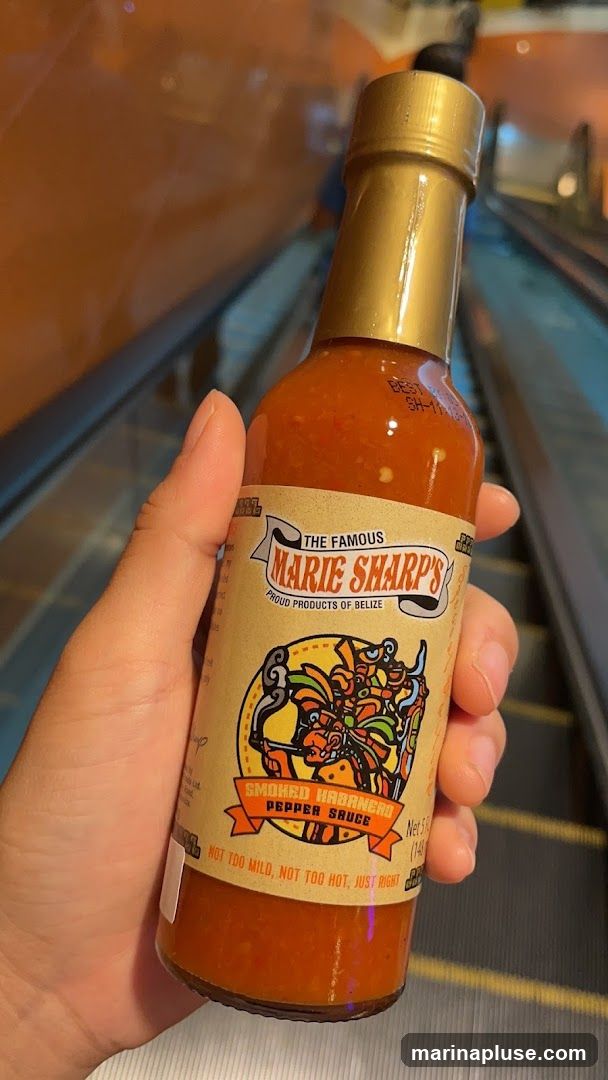 A hand holds a bottle of Marie Sharp's Smoked Habanero Pepper Sauce. The label features the brand name, "Proud Products of Belize," and a graphic illustration. Text on the label reads, "Not too mild, not too hot, just right." The background shows an escalator.