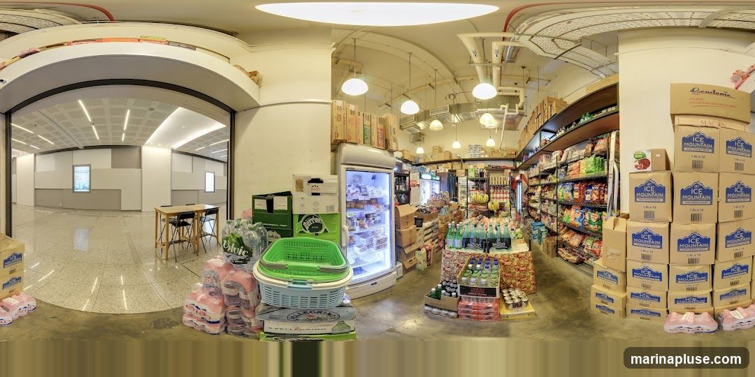A panoramic view of the interior of a grocery store. Shelves are stocked with various food items, snacks, and beverages. A refrigerated display case is filled with products. Stacks of bottled water and other drinks are visible near the entrance. In the background, an empty room with tables and chairs can be seen through an archway.
