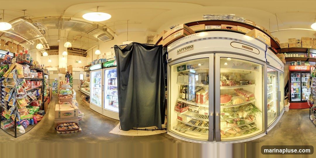 A panoramic view of the interior of a grocery store. Shelves are stocked with various food items, including packaged goods, produce, and refrigerated items. There are also several large refrigerators and freezers visible, some displaying beverages and other products. Boxes are stacked on shelves and on top of some of the refrigerators. A dark curtain hangs in front of one of the refrigerators.