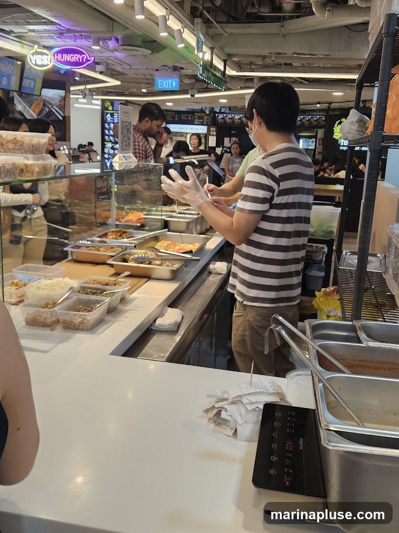 A food stall counter with various dishes displayed in metal trays. A staff member wearing gloves is preparing food. Customers are visible in the background, and a neon sign that says "YES! HUNGRY?" is illuminated above. There are also stacks of receipts on the counter.