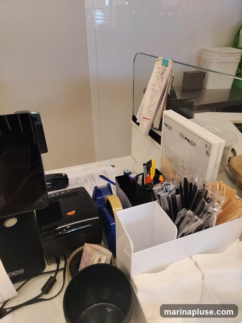 A close-up view of a counter with various supplies, including a receipt printer, tape dispenser, pens, straws, wooden stirrers, and napkins. A black trash can is in the foreground. A stack of receipts is clipped to a glass partition.