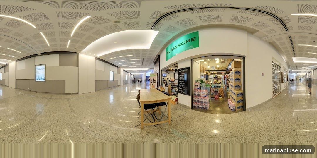 A wide-angle view of a store called AL MARCHE, with its green signage prominently displayed above the entrance. The store appears to be a small grocery or convenience store, with shelves stocked with various products visible through the open doorway. In front of the store, there is a wooden table with chairs, suggesting a small seating area. The store is located within a modern, well-lit indoor walkway or mall.