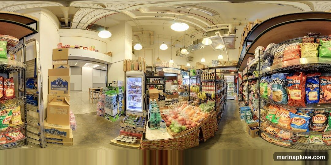 A panoramic view of the interior of a grocery store, showcasing aisles stocked with various products. Shelves are filled with bags of chips, boxes of snacks, bottled water, and canned goods. A refrigerated display case is visible, along with a produce section featuring fruits and vegetables. The store appears well-lit with overhead lighting.