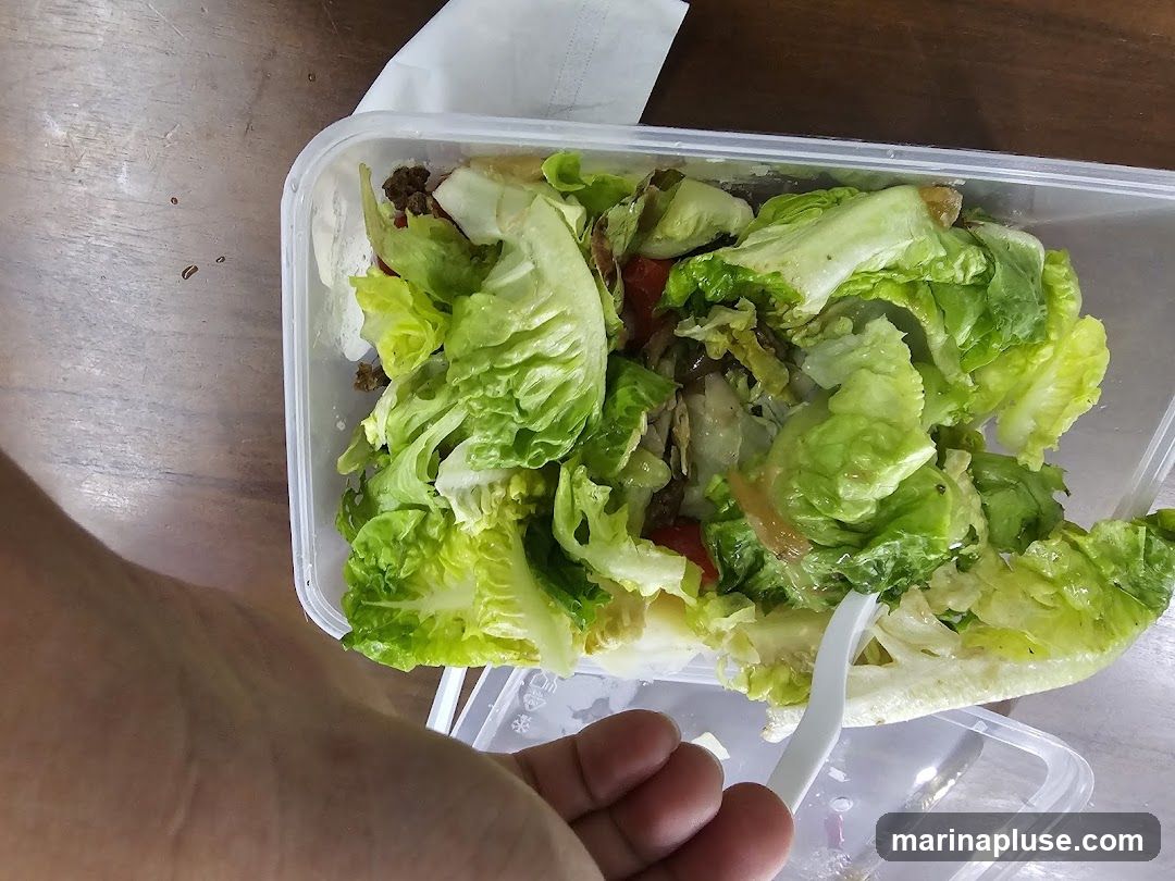 A close-up overhead view of a salad in a clear plastic takeaway container. The salad consists primarily of lettuce leaves, with some visible pieces of tomato and what appears to be ground meat. A white plastic fork is partially submerged in the salad, and a hand is visible in the lower left corner, holding the fork. The container is placed on a wooden surface.