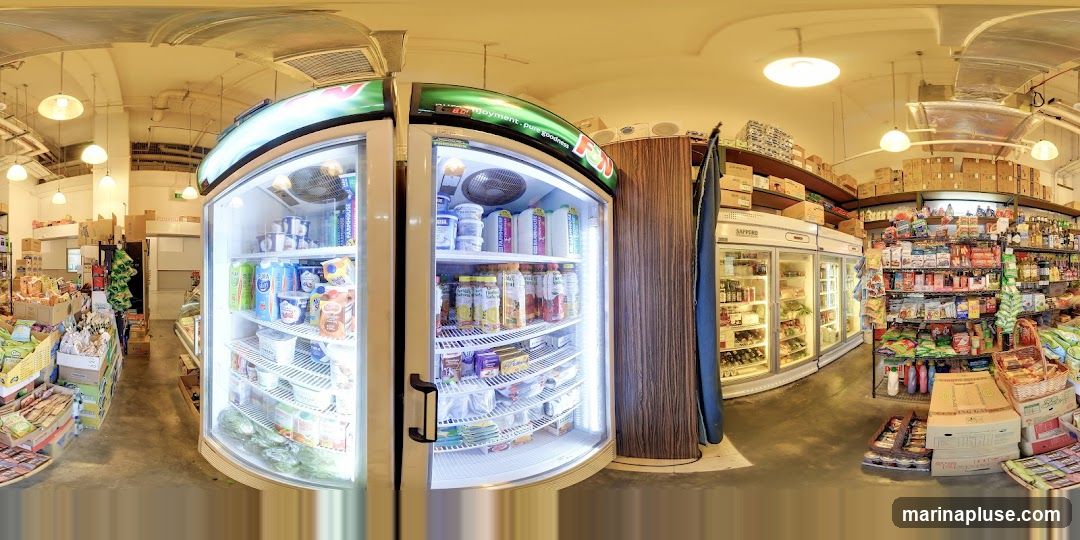 A panoramic view of the interior of a grocery store. In the foreground, there is a large, illuminated refrigerated display case filled with various dairy products and beverages. To the right, shelves are stocked with packaged goods, snacks, and household items. Further back, more refrigerated units and shelves of products are visible, along with stacked boxes on higher shelves. The store appears well-stocked with a variety of items.