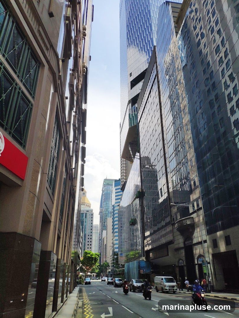 A street scene in a modern city with tall glass and stone buildings on either side. The sky is bright and partly cloudy. Traffic, including cars, motorcycles, and a van, moves along the road. Pedestrians are visible on the sidewalk. The perspective is looking down the street, emphasizing the height of the architecture.