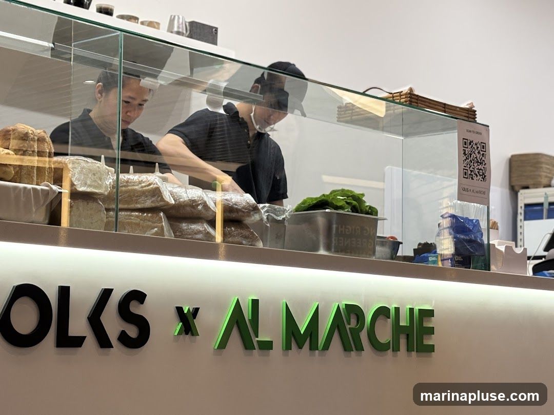 Two men are working behind a counter in a sandwich shop. There are loaves of bread wrapped in plastic on display, as well as a container of lettuce and other ingredients. A QR code for ordering is visible on the glass partition. The shop's name, "FOLKS x AL MARCHE", is displayed prominently on the front of the counter.