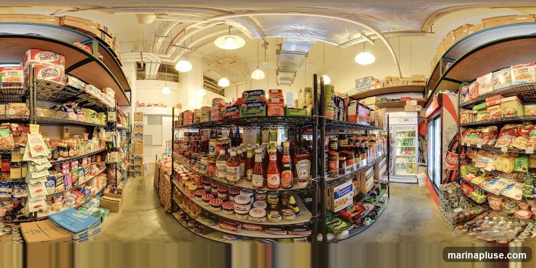 A panoramic view of the interior of a grocery store, showcasing aisles stocked with various food products. Shelves are filled with canned goods, jars of condiments, boxes of cereal, snacks, and beverages. A red and white Coca-Cola branded refrigerator is visible, along with a white upright cooler. The store appears well-organized with a wide selection of items.
