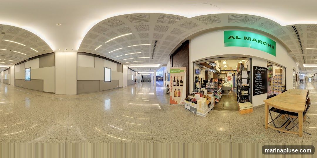 A panoramic view of a shop front with the name AL MARCHE displayed in green lettering above the entrance. The shop appears to be a deli or market, with displays of food items, drinks, and shelves stocked with products. A blackboard menu is visible, listing sandwiches and salads. A wooden table and chairs are set up outside the shop. The shop is located in a modern, well-lit indoor walkway or mall.