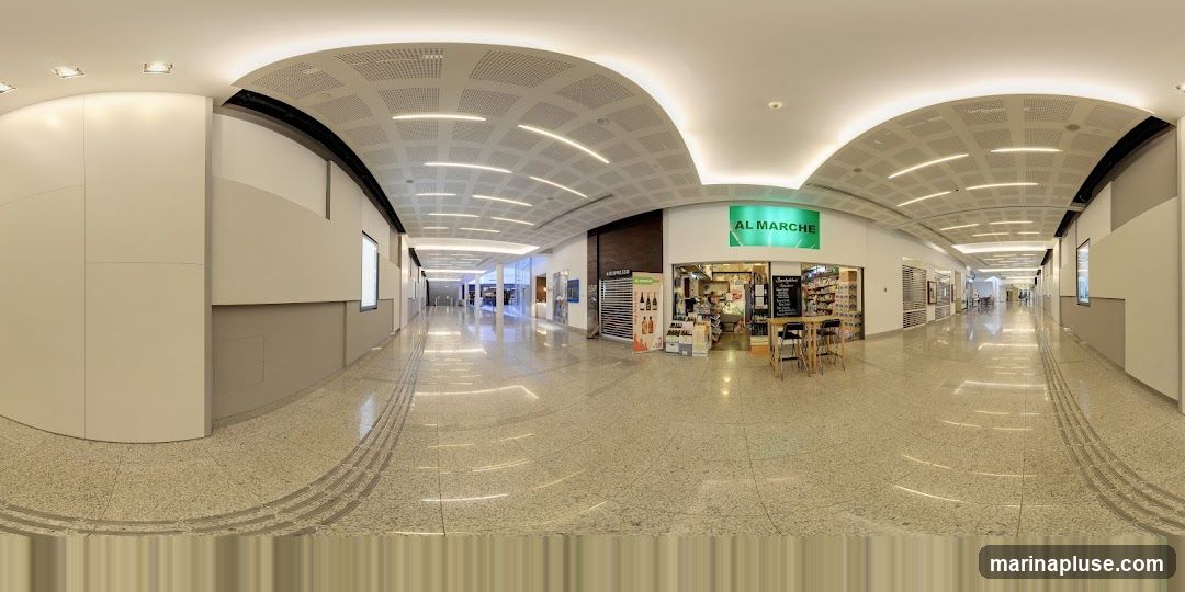 A panoramic view of a shopping mall interior with the AL MARCHE store prominently featured. The store has a green sign with white lettering that reads "AL MARCHE". Inside, shelves are stocked with various products, and there is a counter area. A menu board is visible outside the store, along with tables and chairs. The mall features a polished floor, modern lighting, and a curved ceiling. Other shops and walkways are visible in the background.