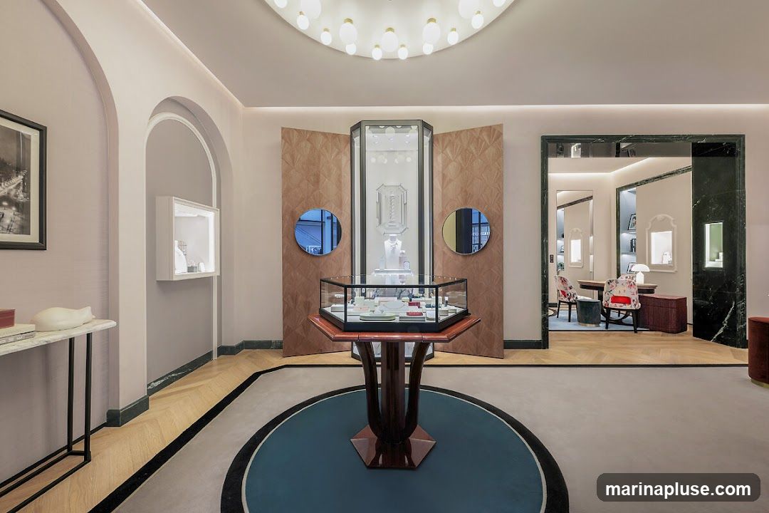 The interior of a luxurious jewelry store features a central display case with various items, a tall display cabinet behind it, and a decorative wall panel. To the left, a console table holds a sculpture and a framed picture. An arched alcove contains a smaller display case. The store has a sophisticated ambiance with soft lighting, wooden floors, and a circular rug. An adjacent room is visible through an open doorway, showing a seating area with chairs and a table.