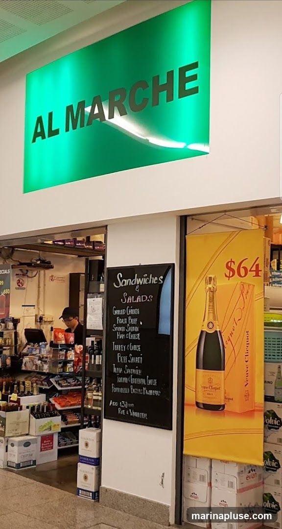 A view of the entrance to a store called Al Marche, with a green illuminated sign above. To the right of the entrance is a yellow banner advertising Veuve Clicquot champagne for $64. A blackboard menu lists various sandwiches and salads, including Grilled Chicken, Roast Beef, Smoked Salmon, Ham & Cheese, Turkey & Cheese, Beef Salad, Tuna Sandwich, and Toronto Continental Cheese. The interior of the store is visible, showing shelves stocked with goods and a person behind a counter.