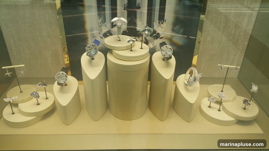 A display window showcases various pieces of jewelry, including watches, earrings, rings, and a bracelet. The jewelry is presented on cream-colored stands of different shapes and sizes, against a backdrop featuring a stylized image of a lighthouse and diamond patterns. Some pieces are designed with intricate details, resembling butterflies and wings.