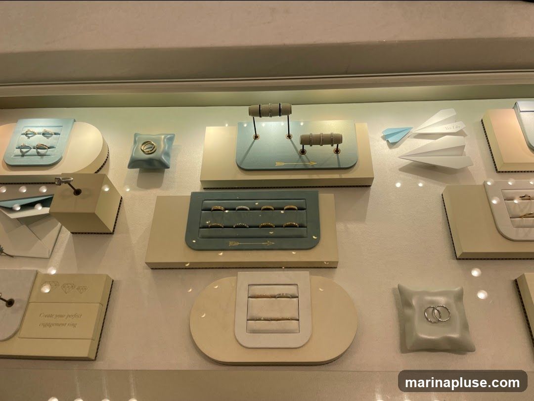 A display case filled with various rings and jewelry. Rings are presented on light blue and cream-colored stands and cushions. Some displays feature multiple rings, while others showcase a single piece. There are also paper airplanes, one with the text "Yes I do" visible. One display has the text "Create your perfect engagement ring" and diamond graphics.