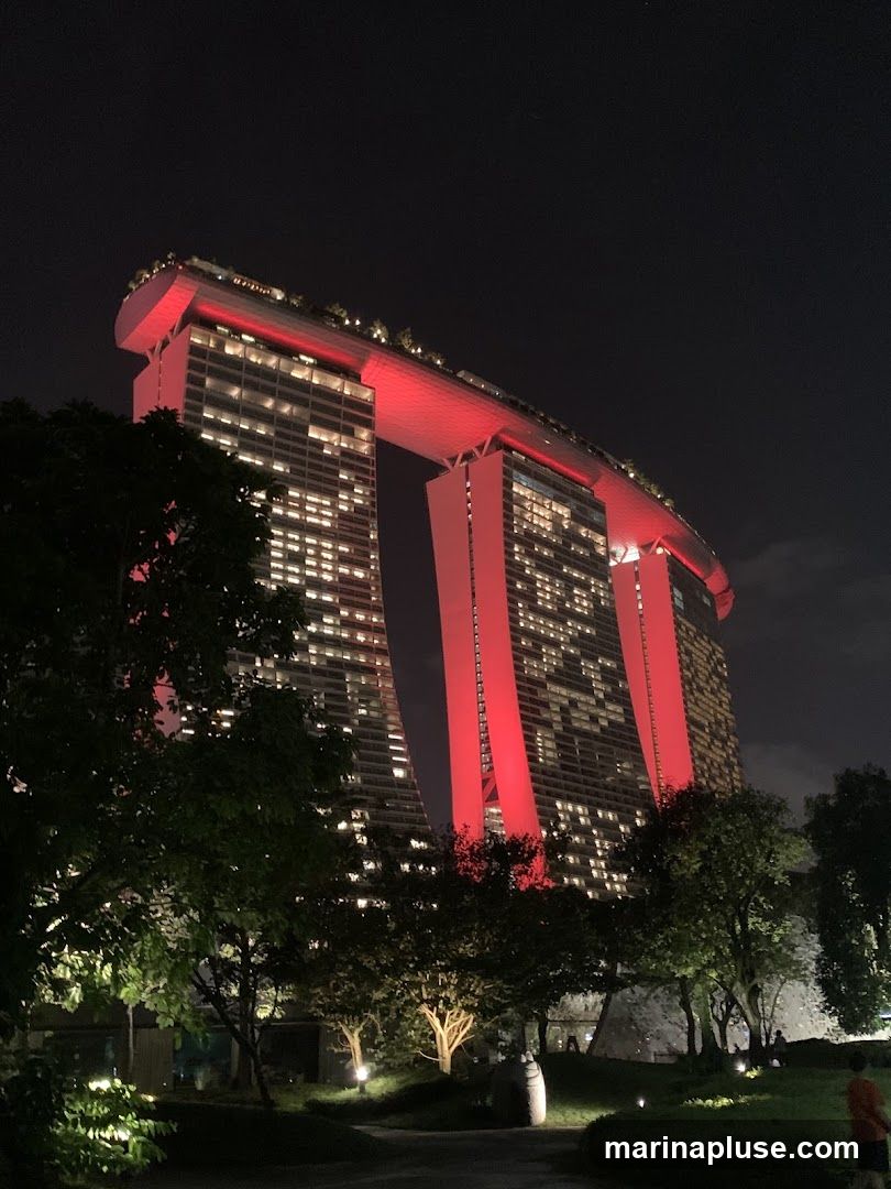 A nighttime view of the Marina Bay Sands hotel in Singapore, illuminated with red lights. The iconic building's three towers are visible, connected by the SkyPark at the top. The foreground features trees and landscaping, with some lights illuminating the park area. A person is visible on the right side of the frame.