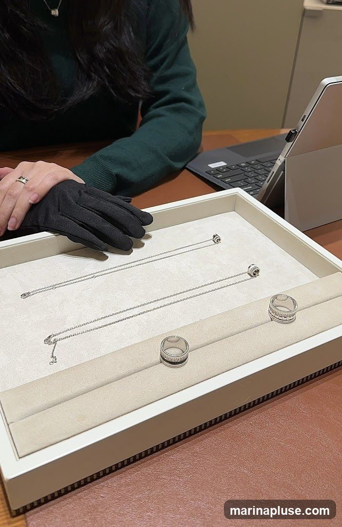 A person wearing black gloves and a ring is seated at a table with a laptop and a display tray of jewelry. The tray contains two necklaces and two rings, all appearing to be made of silver or white gold with small stones.