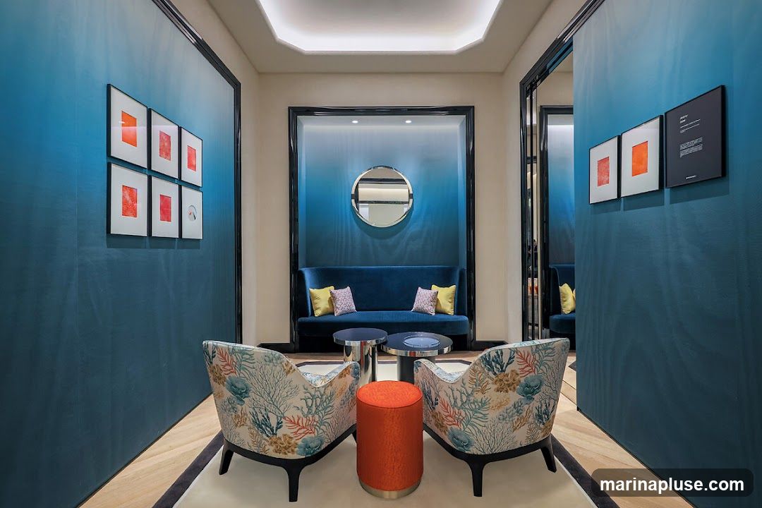A stylish waiting area features deep blue ombre walls with framed artwork. Two armchairs with a coral and floral pattern face a dark blue sofa. In the center, two metallic coffee tables and an orange ottoman create a seating arrangement. A circular mirror hangs above the sofa.