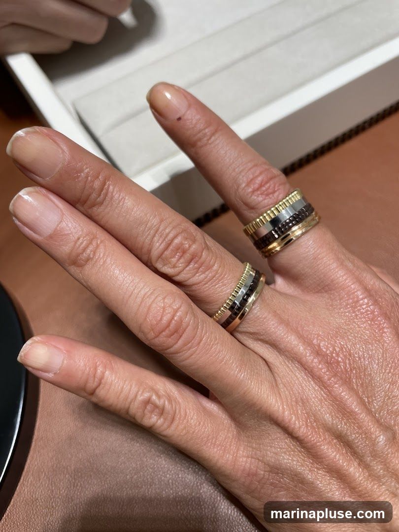 A close-up shot of a person's hand wearing two rings. The rings are made of gold and feature a textured design with dark accents. The background is blurred, with a hint of a jewelry box visible.