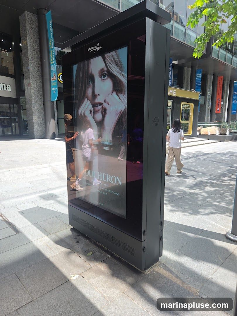 A digital advertisement display stands on a paved walkway. The screen shows a close-up of a woman's face with the text "STELLAR ACE" and "BOUCHERON" below. Several people are walking by in the background. The display is located in an outdoor urban environment with modern architecture visible.
