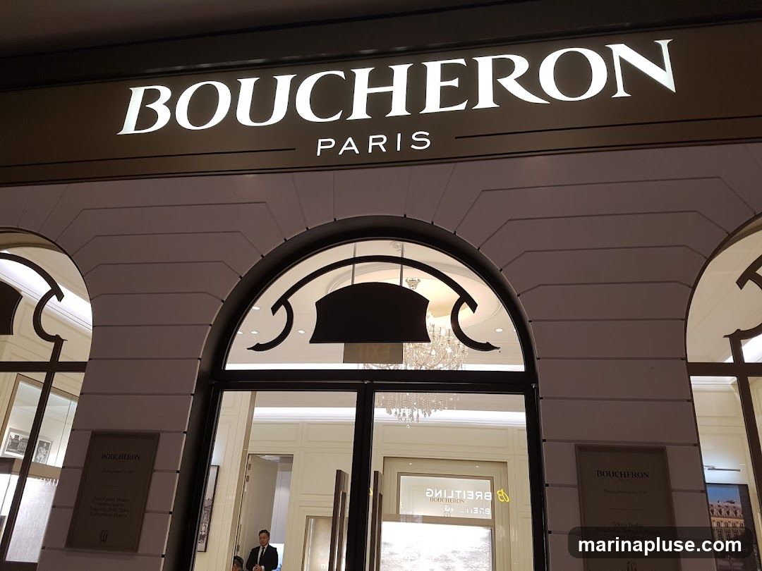 The exterior of a Boucheron Paris jewelry store. The prominent sign above the entrance reads "BOUCHERON PARIS" in large white lettering. The entrance features large glass doors and arched windows, revealing a chandelier and interior displays. Plaques with the Boucheron logo and a list of locations are visible on the facade.