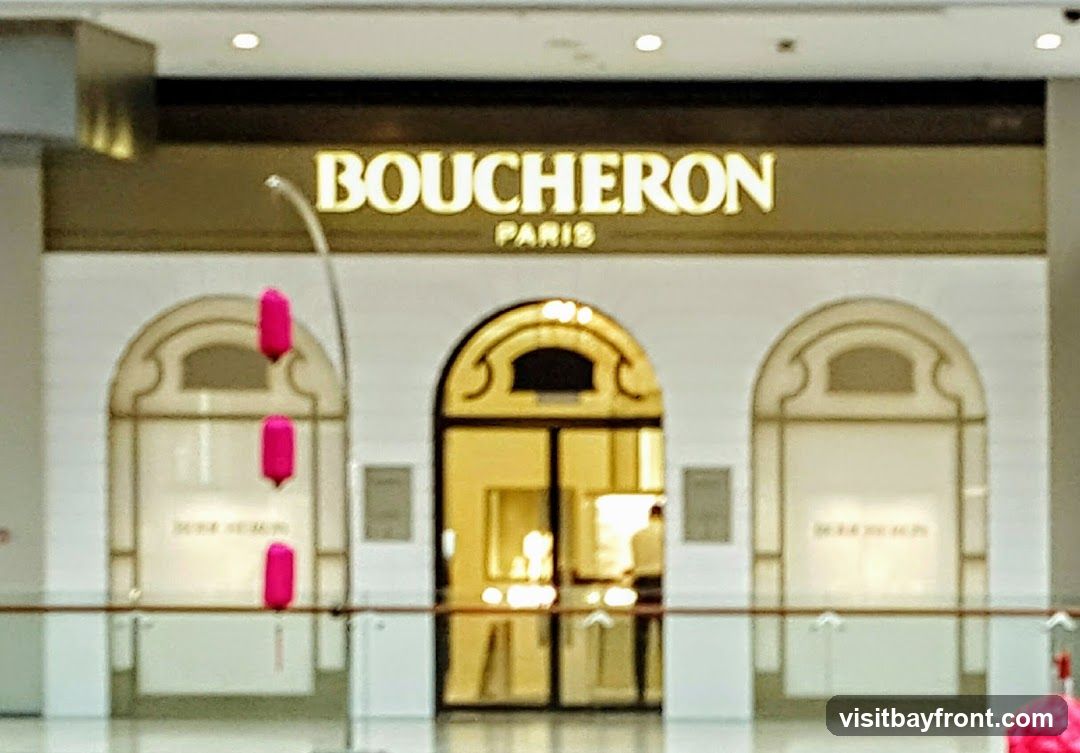 The exterior of a Boucheron Paris jewelry store. The store has a large, arched entrance with double glass doors. Above the entrance, the words "BOUCHERON PARIS" are illuminated in gold lettering. The storefront features decorative arched windows on either side of the entrance. Three bright pink, elongated decorative elements hang from the ceiling in front of the left window.