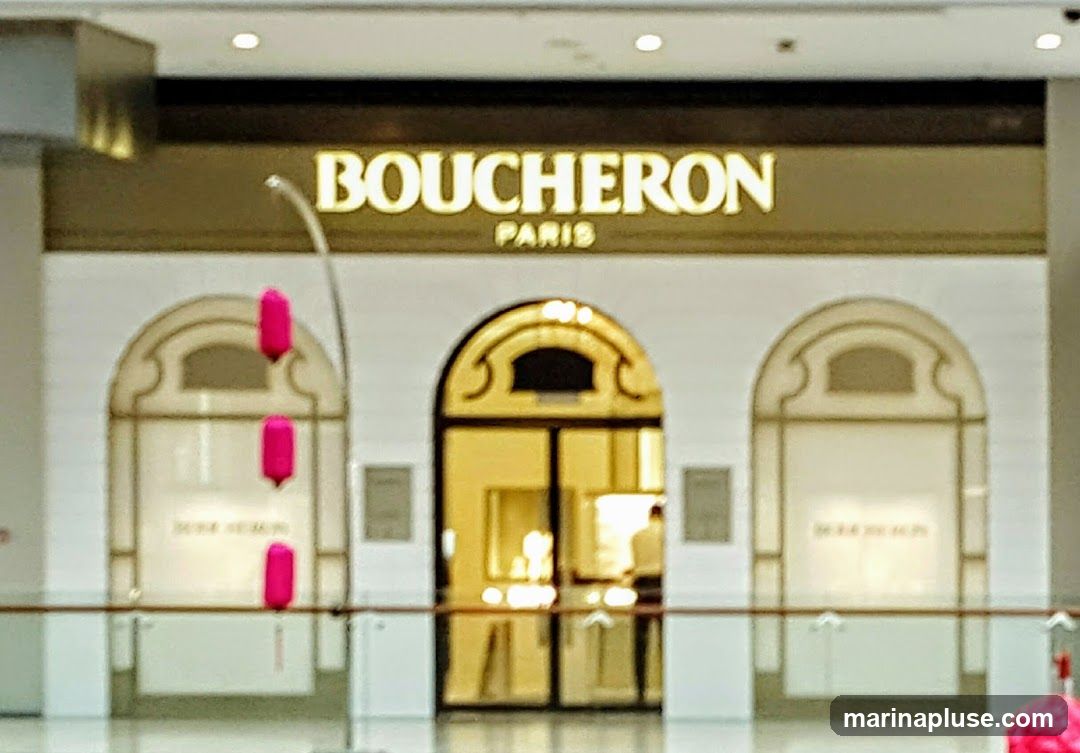 The exterior of a Boucheron Paris jewelry store. The store has a large, arched entrance with double glass doors. Above the entrance, the words "BOUCHERON PARIS" are illuminated in gold lettering. The storefront features decorative arched windows on either side of the entrance. Three bright pink, elongated decorative elements hang from the ceiling in front of the left window.