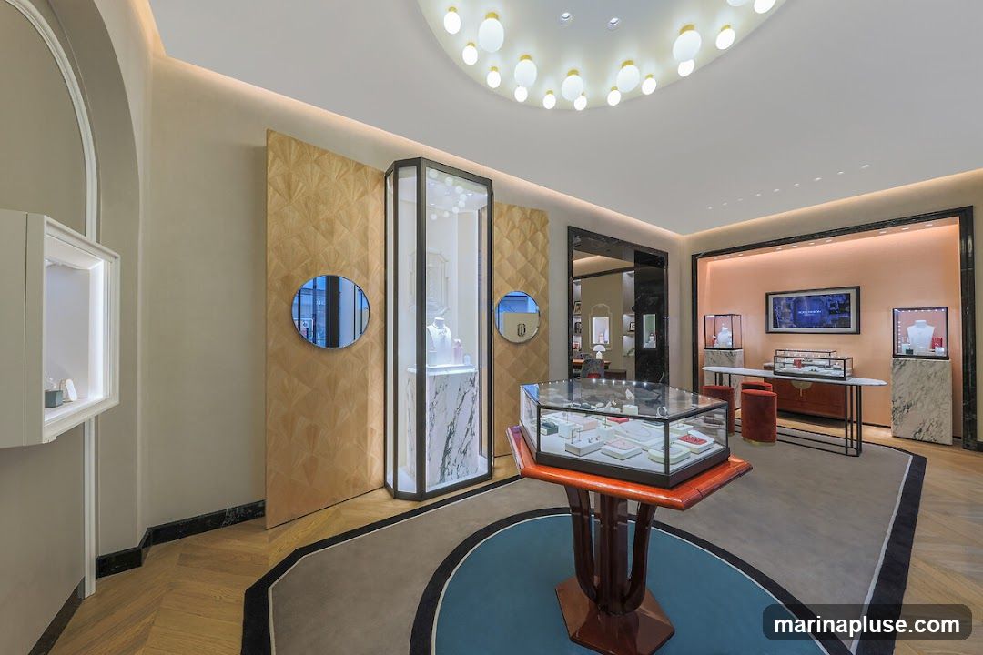 The interior of a high-end jewelry store is showcased, featuring elegant display cases filled with jewelry. The store has a sophisticated design with warm-toned walls, patterned wall panels, and a unique ceiling light fixture. Mirrors are strategically placed, and the flooring is a combination of wood and carpet. In the background, a digital display shows an advertisement, and more display cases are visible, along with seating.