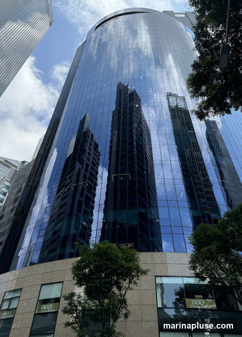 A low-angle shot of a modern glass-facade skyscraper reflecting the sky and surrounding buildings. The lower section of the building features windows with visible signage for "BDO Private Banking". Trees are visible in the foreground.