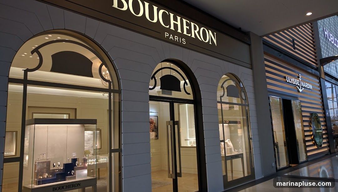 The exterior of a Boucheron Paris jewelry store, with large arched windows displaying jewelry in glass cases. The store's name and 'Paris' are visible in illuminated lettering above the entrance. To the right, the storefront of Ulysse Nardin is visible.