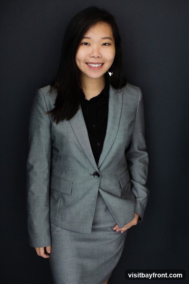A young Asian woman wearing a grey suit jacket and skirt over a black collared shirt smiles at the camera. She has long, dark hair and is wearing triangular earrings. The background is a plain dark grey.