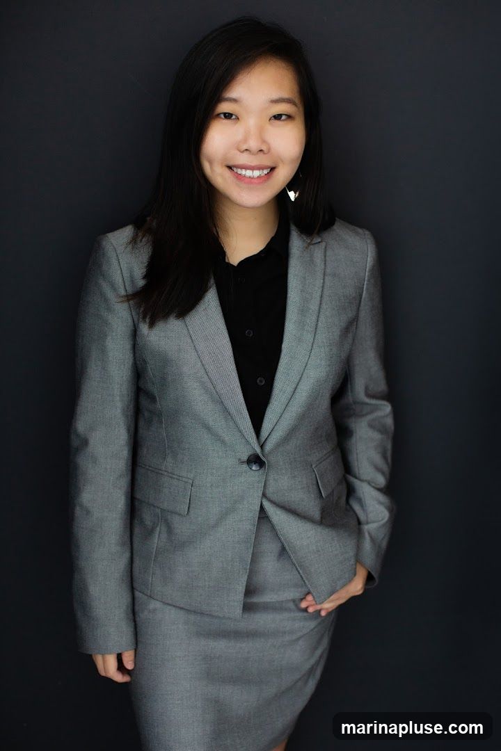 A young Asian woman wearing a grey suit jacket and skirt over a black collared shirt smiles at the camera. She has long, dark hair and is wearing triangular earrings. The background is a plain dark grey.