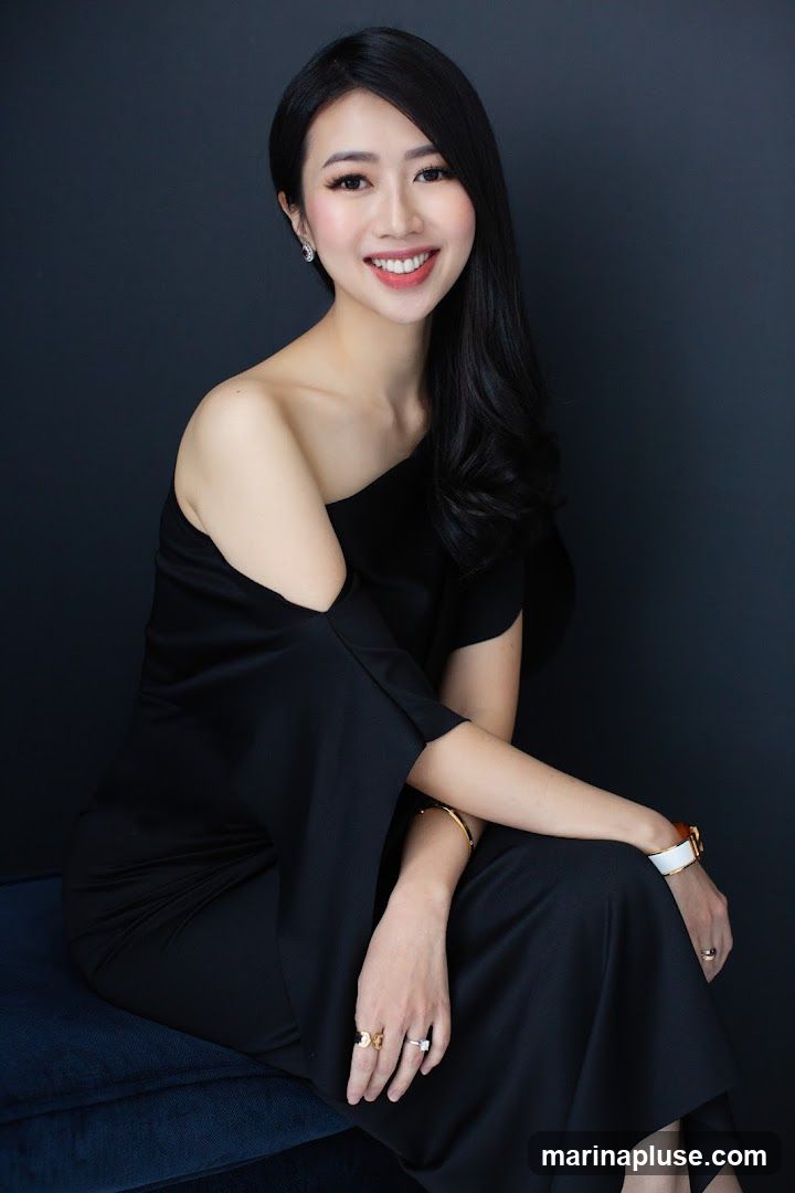 A young Asian woman with long, dark, wavy hair is smiling and looking at the camera. She is wearing a black off-the-shoulder dress and several rings and bracelets. She is seated on a dark blue cushion against a dark background.