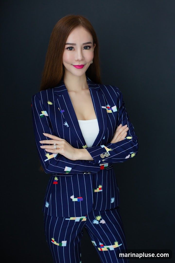 A woman with long, brown hair and full makeup poses with her arms crossed. She is wearing a navy blue pinstripe suit with a colorful, abstract pattern. Underneath the suit jacket, she wears a white top. She has rings on her fingers. The background is a plain dark gray.