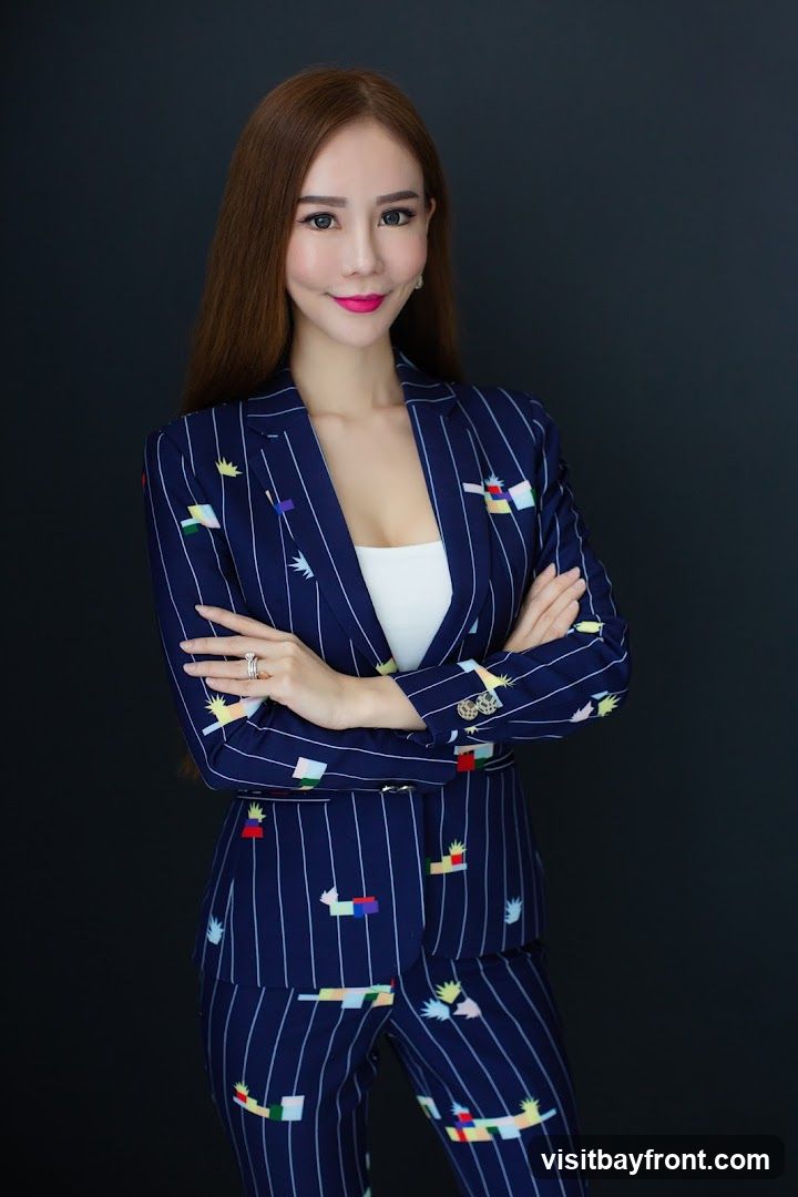 A woman with long, brown hair and full makeup poses with her arms crossed. She is wearing a navy blue pinstripe suit with a colorful, abstract pattern. Underneath the suit jacket, she wears a white top. She has rings on her fingers. The background is a plain dark gray.
