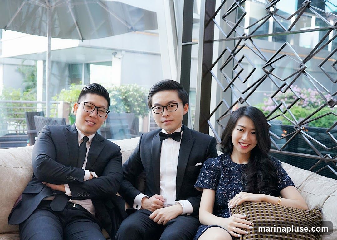 Three people, two men and one woman, are sitting on a plush couch. The two men are wearing black suits and ties, with one man also wearing a tuxedo with a bow tie. The woman is wearing a dark blue lace dress. They are all smiling and looking towards the camera. The background appears to be an indoor setting with large windows, greenery visible outside, and a decorative metal screen.
