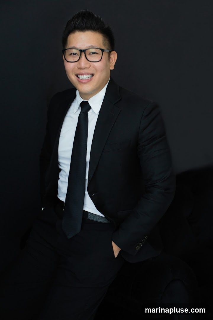 A portrait of a smiling East Asian man wearing a black suit, white shirt, and black tie. He has black-rimmed glasses and his hair is styled upwards. He is leaning against a dark surface with his hands in his pockets. The background is a solid dark gray.