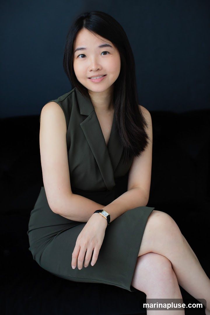 A portrait of a woman with long dark hair, wearing a sleeveless green dress and a watch on her left wrist. She is sitting with her legs crossed against a dark background.