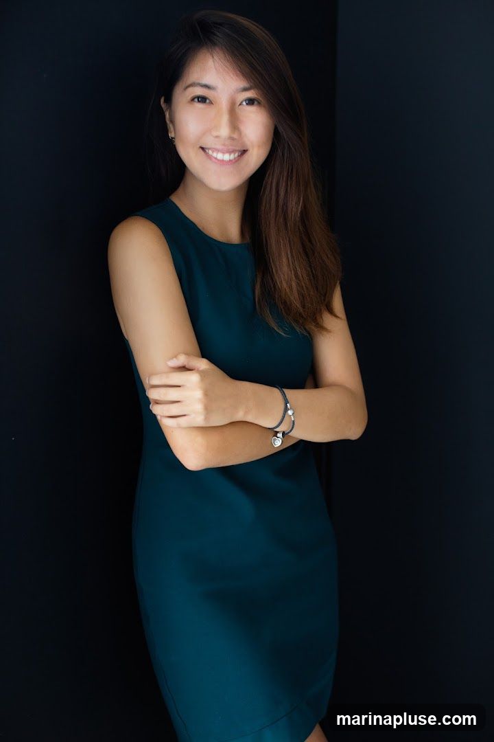 A young Asian woman with long, dark hair smiles at the camera. She is wearing a sleeveless, dark teal dress and a bracelet on her left wrist. Her arms are crossed in front of her. The background is a solid black.