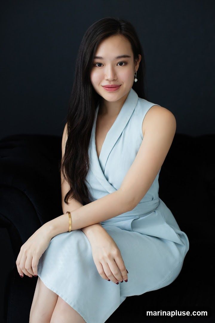 A young woman with long, dark hair is seated on a dark couch against a dark background. She is wearing a light blue sleeveless wrap dress and a gold bracelet on her left wrist. She is also wearing pearl earrings. She is looking at the camera with a slight smile.