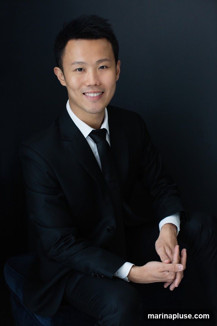 A portrait of a man wearing a black suit, white shirt, and black tie. He is smiling and looking at the camera. The background is dark.