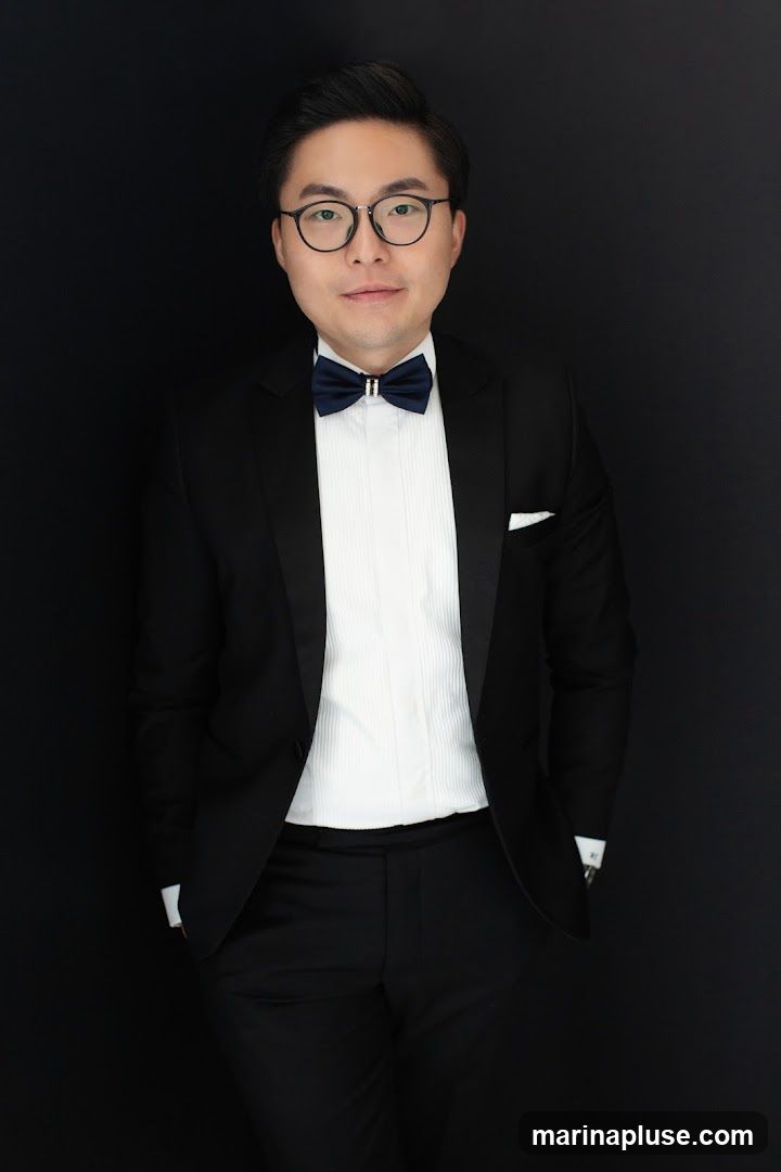 A man wearing a black tuxedo with a white pleated shirt and a navy blue bow tie stands against a black background. He has on round glasses and his hands are in his pockets.