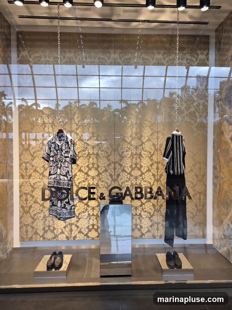A store window display featuring clothing and shoes from Dolce & Gabbana. The background is a gold damask pattern. On the left, a short-sleeved patterned shirt is displayed. On the right, a striped long-sleeved top is shown with black pants. Two pairs of black shoes are displayed on platforms at the bottom of the window. The Dolce & Gabbana logo is prominently displayed in the center.