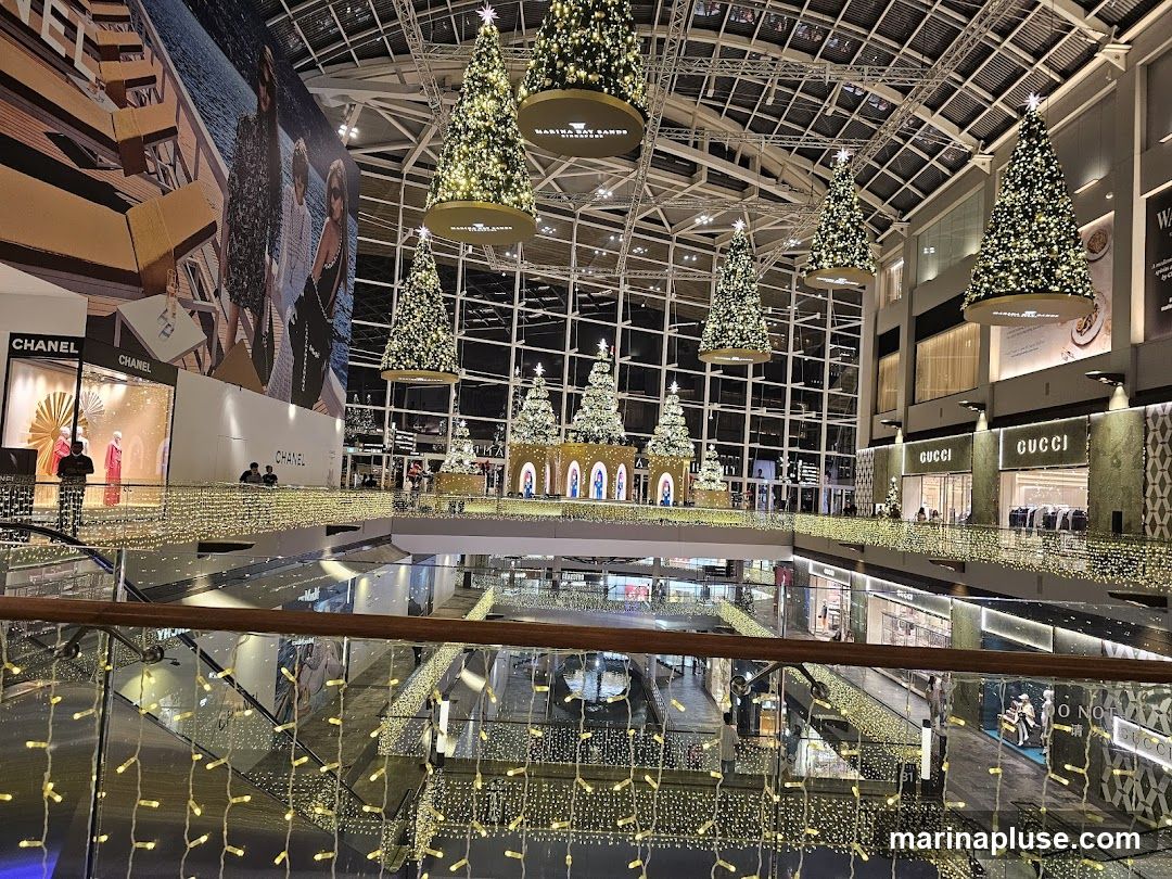 A wide shot of a shopping mall interior decorated for Christmas. Several large Christmas trees are suspended from the ceiling, adorned with lights and ornaments. Smaller Christmas trees are displayed on a lower level. The mall features various luxury brand stores, including Chanel and Gucci, with their prominent signage visible. The railings and walkways are decorated with strings of warm white fairy lights, creating a festive atmosphere. A large mural is visible on one wall.