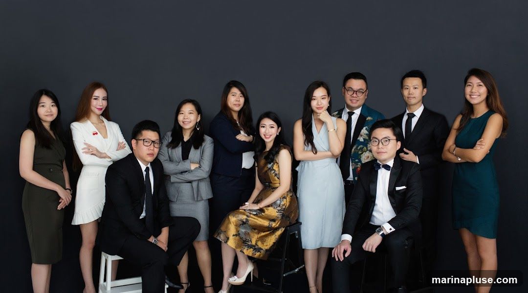 A diverse group of eleven individuals, appearing to be business professionals, are posing for a studio portrait against a dark grey backdrop. The group consists of six men and five women, all dressed in formal attire. The men are wearing suits and ties, with one man in a tuxedo and another in a suit with a floral pattern. The women are wearing dresses and suits. The individuals are arranged in two rows, with some standing and others seated. The overall impression is one of professionalism and teamwork.