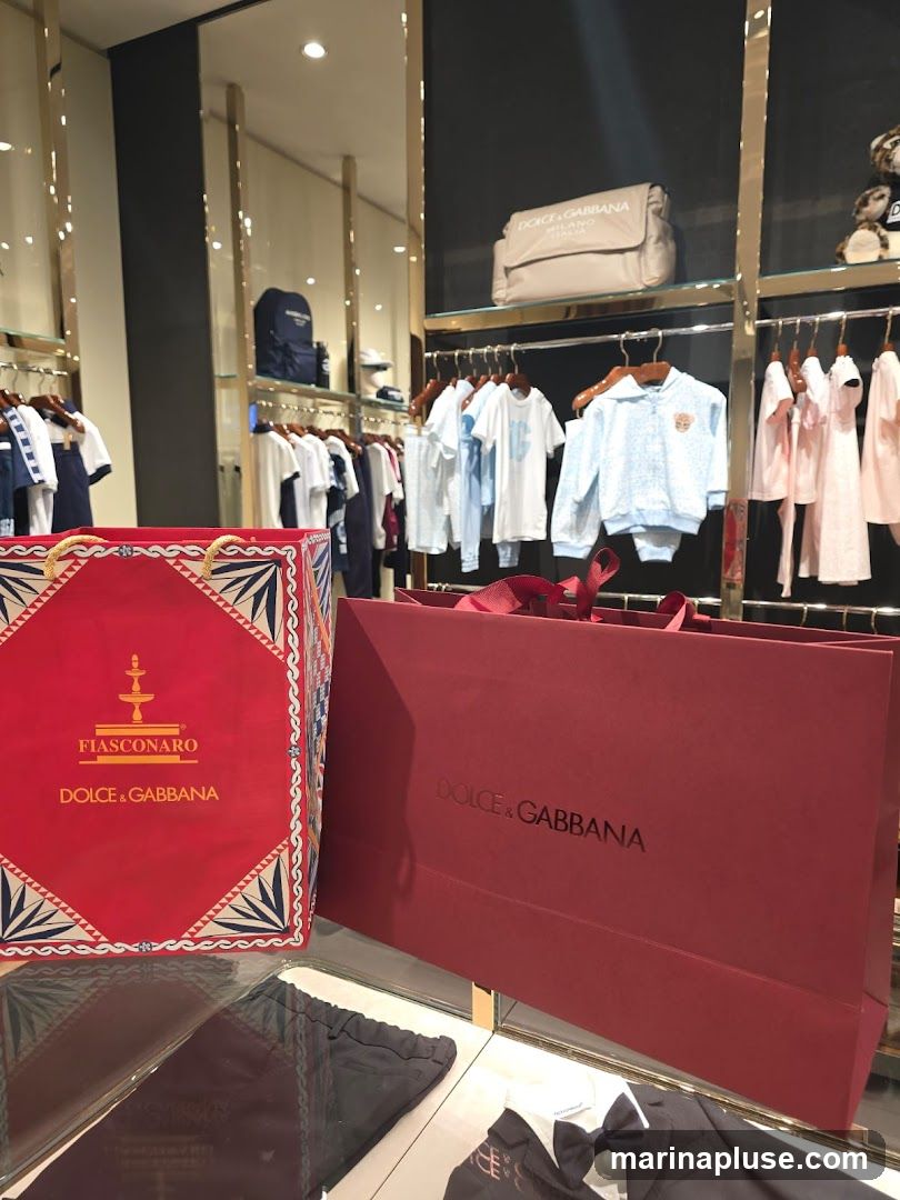 A retail display featuring children's clothing and branded shopping bags. In the foreground, a red box with gold trim and the "FIASCONARO DOLCE & GABBANA" logo is visible, alongside a large red Dolce & Gabbana shopping bag. Behind these, racks of children's apparel, including t-shirts and outfits in light blue and pink, are displayed. A beige diaper bag with the "DOLCE & GABBANA MILANO" logo is on a shelf above the clothing racks.